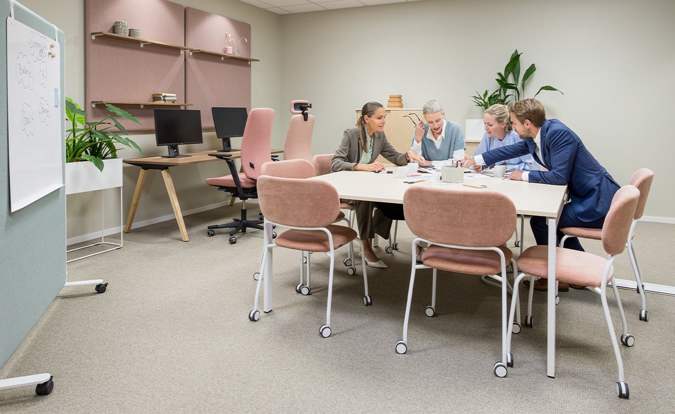 A meeting room with a large white meeting table surrounded by pink and beige upholstered chairs with wheels. Four people are engaged in a discussion at the table. In the background, desks with computers, plants, and sound-absorbing panels on the wall can be seen.